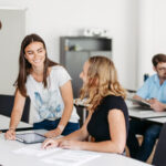 Mehrere Studierende sitzen in einem Klassenraum an Tischen und besprechen sich